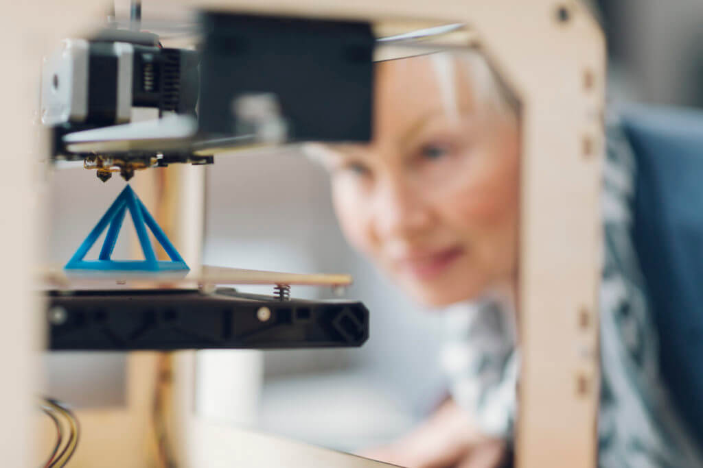 Mature smiling woman working by 3d printer. Selective focus to object in the 3D printer. She is starting printing process for her new 3d project.