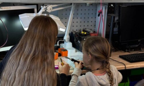 Eine erwachsene Person unterstützt ein Mädchen an einem Werktisch in einer Technikwerkstatt beim Löten einer kleinen elektronischen Schaltung. Beide sitzen nebeneinander unter einer beleuchteten Lupe und halten Werkzeuge in der Hand. Auf dem Tisch stehen Messgeräte, Bauteile und Aufbewahrungsboxen; im Hintergrund sind Regale mit Werkzeugen und grünen Sortierkästen zu sehen.