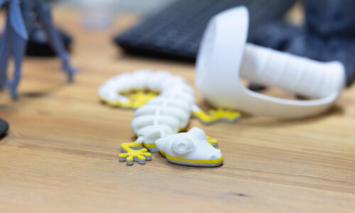 Ein weiß-gelber 3D-gedruckter Gecko liegt auf einem Holztisch, im Hintergrund sind weitere 3D-Druck-Objekte zu erkennen.