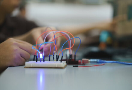 Nahaufnahme eines Elektronik-Experiments mit Steckplatine, bunten Kabeln und leuchtender LED – im Hintergrund sind Hände bei der Arbeit zu sehen.