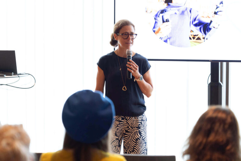 Eine Frau hält ein Mikrofon und spricht auf der Bühne des EdTech Austria Summit 2025. Im Hintergrund ist ein Bildschirm mit einer Präsentationsfolie zu sehen, im Vordergrund das Publikum.