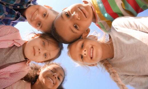 Fünf Kinder stehen im Kreis und schauen lachend in die Kamera, die von unten nach oben fotografiert – der blaue Himmel bildet den Hintergrund.