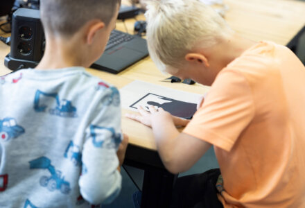 Zwei Kinder arbeiten konzentriert an einem Tisch mit einer schwarzen Plotterfolie. Eines der Kinder löst gerade Teile aus dem Motiv.