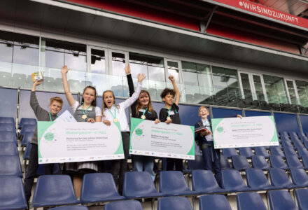 Sechs Kinder jubeln mit Urkunden und Plakaten bei der Preisverleihung der MINT Challenge 2024 im Stadion.
