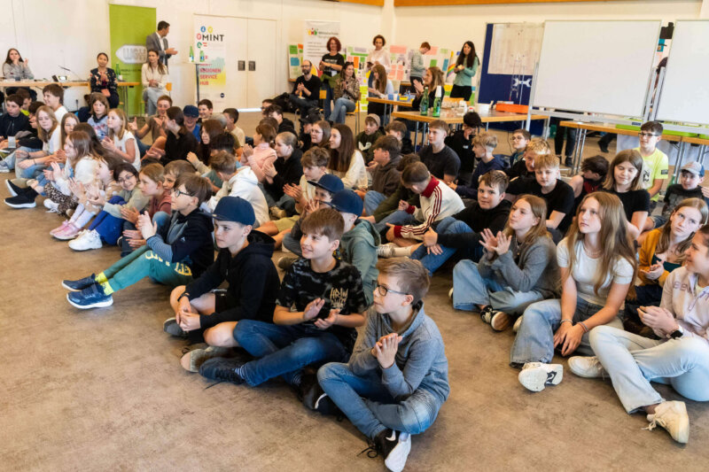 Gruppe von Schüler:innen sitzt bei der MINT-Fair 2025 am Boden und applaudiert während einer Präsentation.