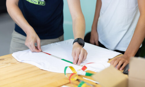 Zwei Personen messen ein weißes T-Shirt mit einem Maßband auf einem Holztisch ab.