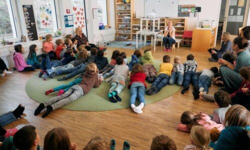 In einem Klassenraum sitzen und liegen zahlreiche Kinder in einem großen Halbkreis auf einem Teppich und hören aufmerksam einer Frau zu, die vorne auf einem Stuhl sitzt und spricht.