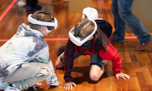 Zwei Kinder tragen Virtual-Reality-Brillen und bewegen sich am Boden eines Raums mit Holzfußboden.