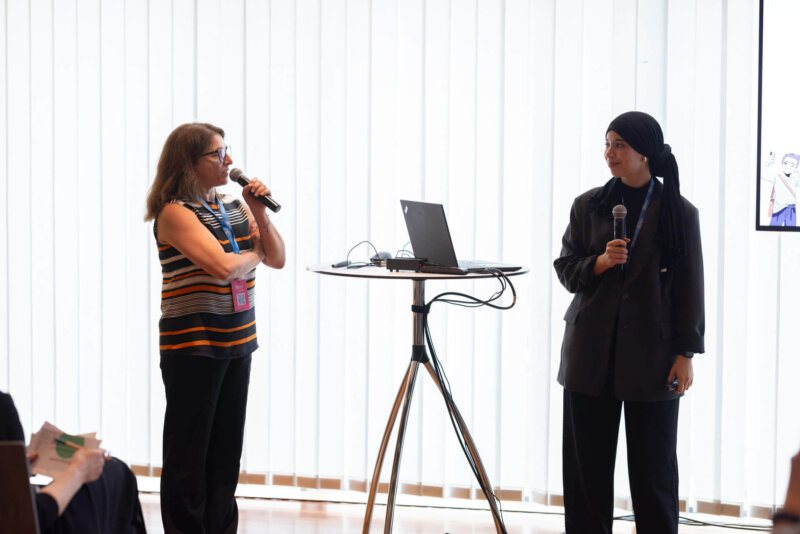 Zwei Frauen stehen auf der Bühne und sprechen beim EdTech Austria Summit 2025. Beide halten ein Mikrofon, zwischen ihnen steht ein Tisch mit einem aufgeklappten Laptop.