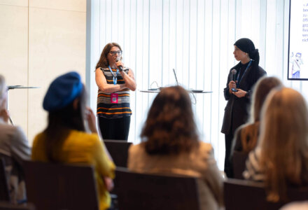 Zwei Frauen stehen auf der Bühne und sprechen beim EdTech Austria Summit 2025. Beide halten ein Mikrofon, zwischen ihnen steht ein Tisch mit einem aufgeklappten Laptop. Im Vordergrund sind die Hinterköpfe des Publikums zu sehen.
