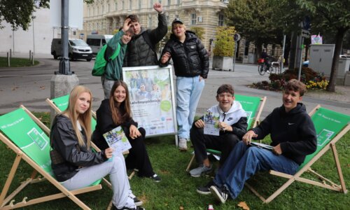 Jugendliche sitzen in einem Park auf Liegestühlen im Halbkreis und blicken in die Kamera. Dahinter ist ein Aufsteller mit der Schrift 
