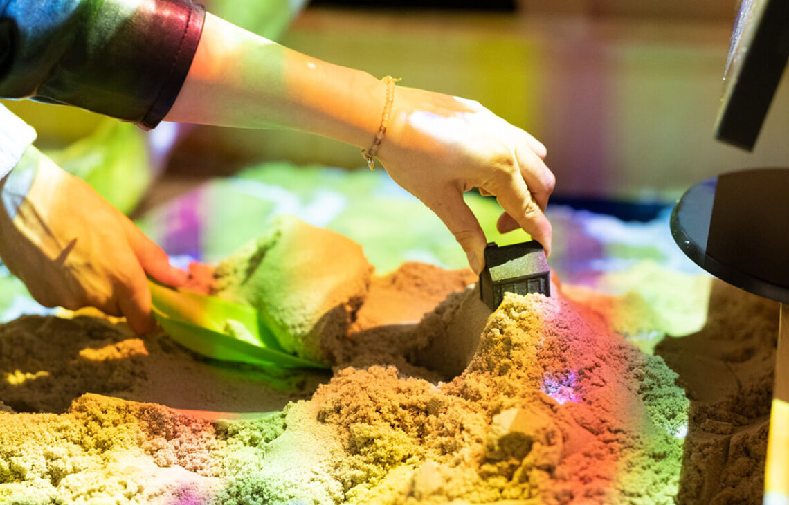 Zwei Hände formen mit Schaufel und Modellhaus eine Landschaft in einem interaktiven Sandkasten, der mit farbigen Höhenlinien projiziert wird.