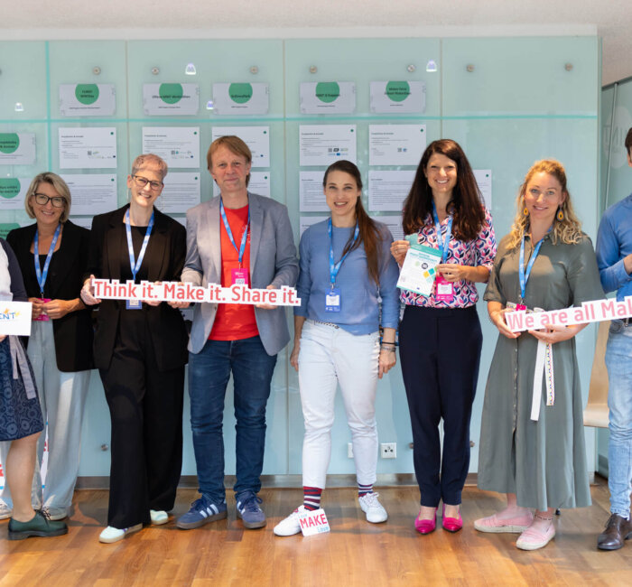 Acht Personen stehen nebeneinander vor einer Wand mit Plakaten und lächeln in die Kamera. Sie tragen Namensschilder und halten Schilder mit Aufschriften wie „Think it. Make it. Share it.“ und „We are all Makers“.