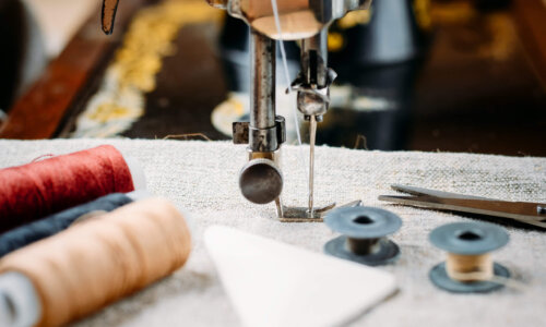 Nahaufnahme einer Nähmaschine mit Stoff unter der Nadel, umgeben von Garnrollen, Spulen, einer kleinen Schere und Schneiderkreide