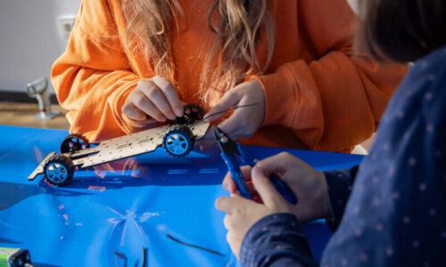 Zwei Mädchen sitzen an einem Tisch und arbeiten gemeinsam an einer kleinen Holzplatte mit vier Rädern. Ein Mädchen hält die Platte, das andere Mädchen hält eine Zange.