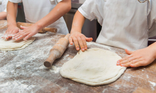 Zwei Kinder formen mit ihren Händen jeweils einen großen runden Teig an einem Tisch. Der Tisch ist voller Mehr und eine kleine Walze liegt auch darauf.