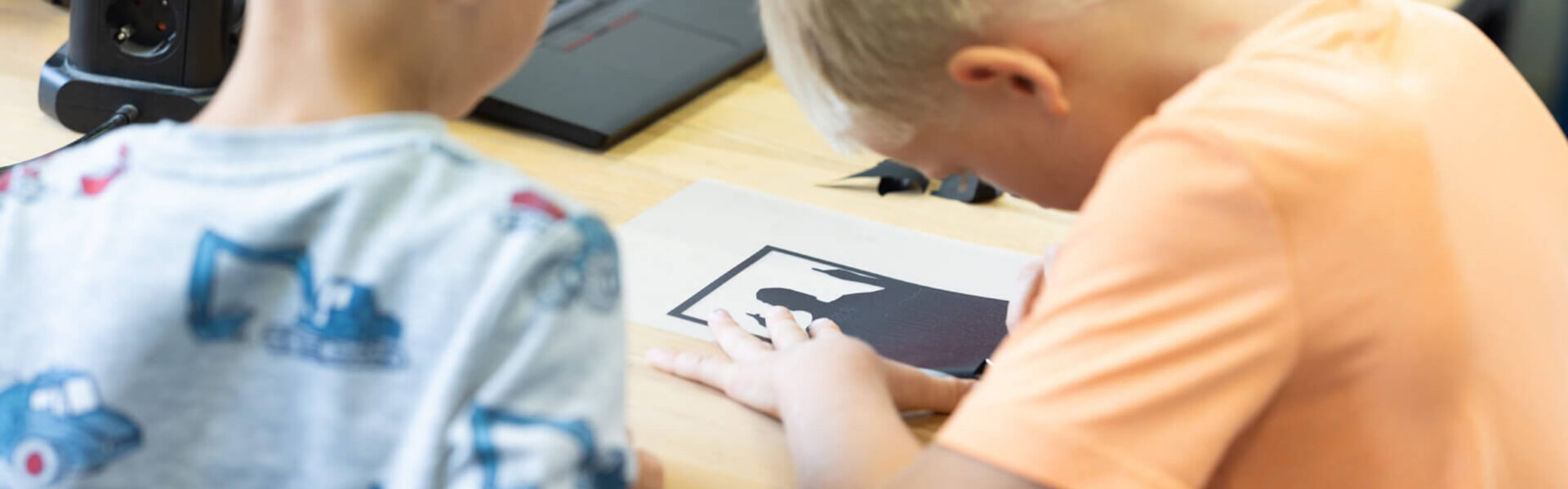 Zwei Kinder arbeiten konzentriert an einem Tisch mit einer schwarzen Plotterfolie. Eines der Kinder löst gerade Teile aus dem Motiv.