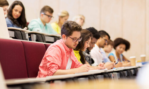 Eine Gruppe Studierender sitzt im Hörsaal und schreibt in ihre Notizbücher am Tisch vor ihnen.