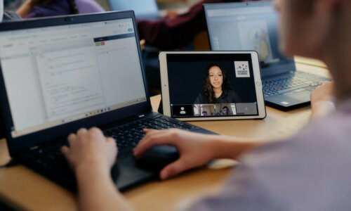 Person arbeitet an einem Laptop, während daneben ein Tablet eine Online-Unterrichtssitzung mit mehreren Teilnehmenden in einer Videokonferenz zeigt; im Hintergrund weitere Laptops in einer Lernumgebung.