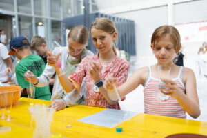 Kinder und Jugendliche führen an einer Mitmachstation der MINT Challenge 2026 einfache Experimente mit farbigen Flüssigkeiten durch und beobachten die Ergebnisse in Bechern und Reagenzgefäßen.