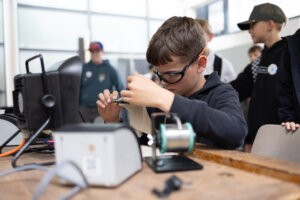 Ein Schüler arbeitet an einer Mitmachstation mit elektronischen Bauteilen und Werkzeugen. Er trägt eine Schutzbrille und konzentriert sich auf eine technische Aufgabe, während weitere Jugendliche die Station beobachten.