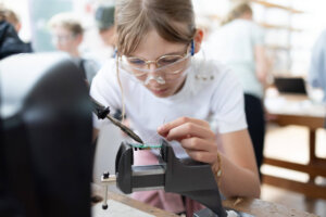 Ein Mädchen trägt eine Schutzbrille und lötet an einer elektronischen Platine. Die Schülerin arbeitet konzentriert an einer Mitmachstation und verbindet Bauteile mit einem Lötkolben.