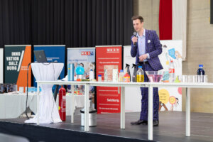 Der Moderator der MINT Challenge vor dem Tisch der Science Show. Auf dem Tisch stehen Utensilien für chemische Experimente.