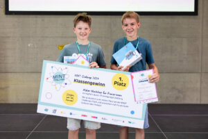 Zwei Schüler der Climate Crew präsentieren ihren ersten Platz bei der MINT Challenge 2026 mit Urkunde, Sachpreisen und Gewinner-Scheck auf der Bühne.