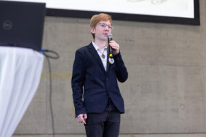 Ein Schüler steht mit Mikrofon auf der Bühne und präsentiert sein Projekt vor Publikum bei der MINT Challenge 2026. Im Hintergrund ist die Projektionsfläche der Veranstaltung zu sehen.