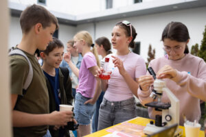 Schülerinnen präsentieren ihr Projekt am Marktstand der MINT Challenge 2026. Eine Schülerin untersucht eine Probe mit einem Mikroskop, während andere Teilnehmende das Experiment beobachten und diskutieren.