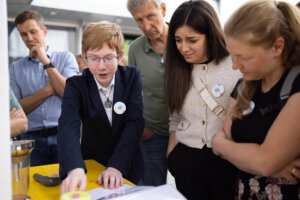 Ein Schüler erklärt Besucher:innen sein Projekt am Marktstand der MINT Challenge 2026. Mehrere Personen verfolgen aufmerksam die Vorführung und diskutieren die Ergebnisse.