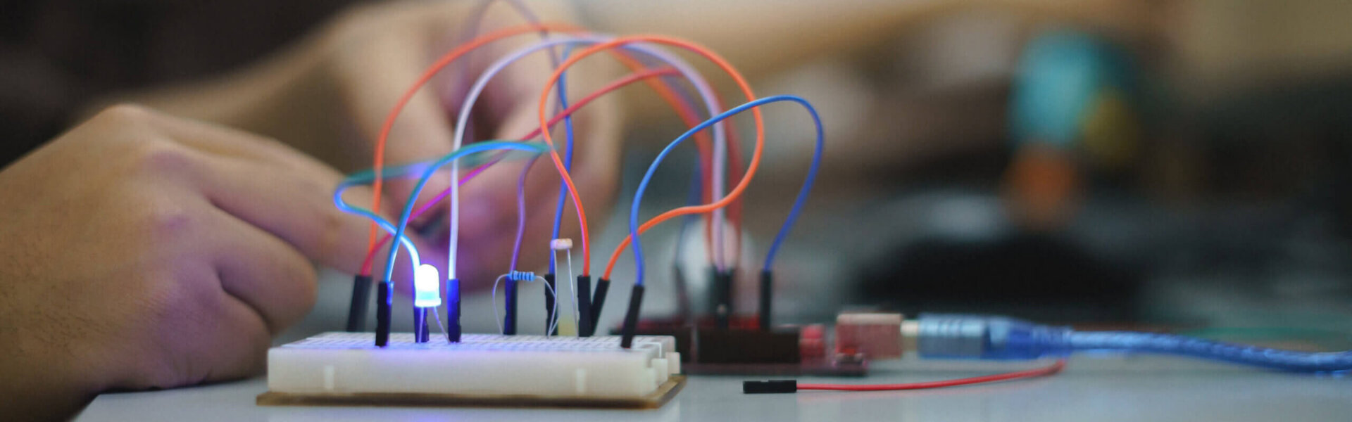 Nahaufnahme eines Elektronik-Experiments mit Steckplatine, bunten Kabeln und leuchtender LED – im Hintergrund sind Hände bei der Arbeit zu sehen.