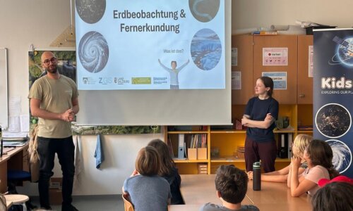 Zwei Referierende präsentieren vor einer Schulklasse das Thema ‚Erdbeobachtung und Fernerkundung‘. Auf der Leinwand ist eine Präsentationsfolie mit Bildern der Erde und der Überschrift ‚Erdbeobachtung & Fernerkundung‘ zu sehen, während die Schülerinnen und Schüler aufmerksam zuhören.