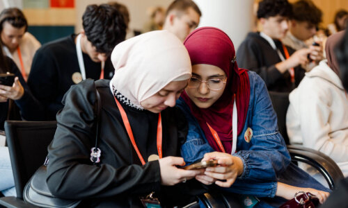 Zwei Teilnehmerinnen sitzen bei einer Veranstaltung nebeneinander und betrachten gemeinsam ein Smartphone. Im Hintergrund arbeiten weitere Jugendliche in einem Veranstaltungsraum.